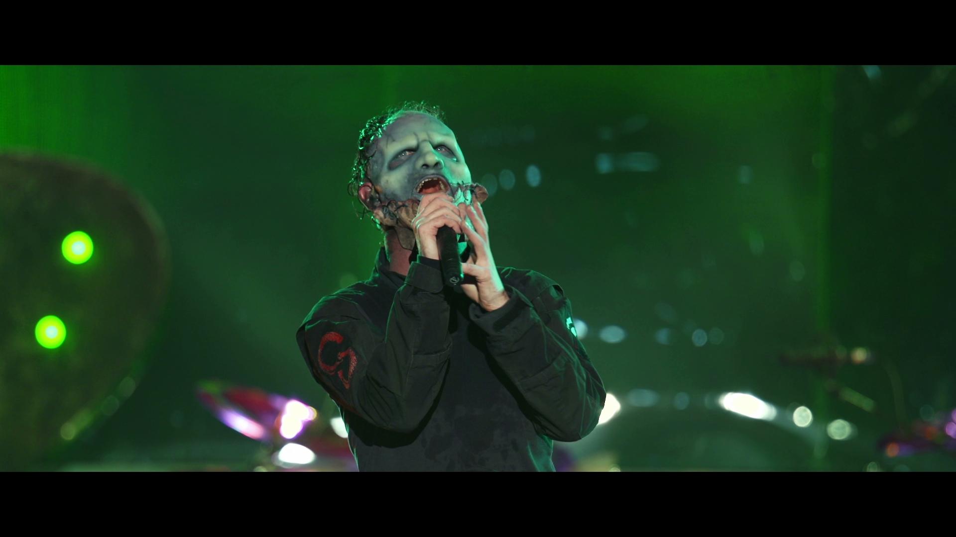 Slipknot frontman wearing theatrical makeup and a black outfit sings into a microphone on a stage with green lighting.