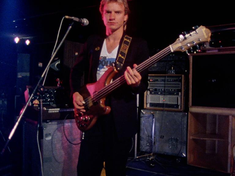 The Police frontman Sting on stage holding a bass guitar, with musical equipment and amplifiers in the background. He is positioned in front of a microphone.