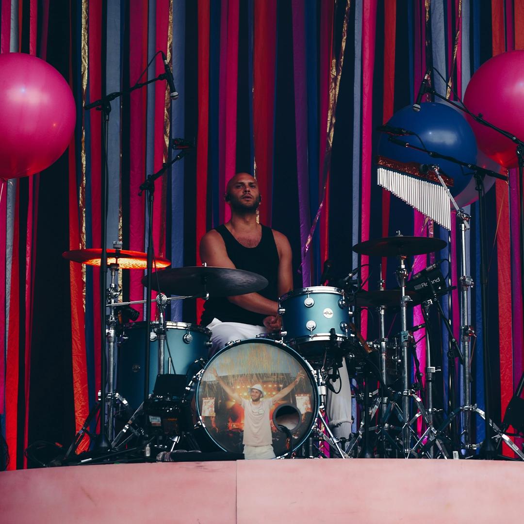 Bombay Bicycle Club drummer sits behind a drum kit, surrounded by colorful hanging ribbons and large pink balloons on a stage.