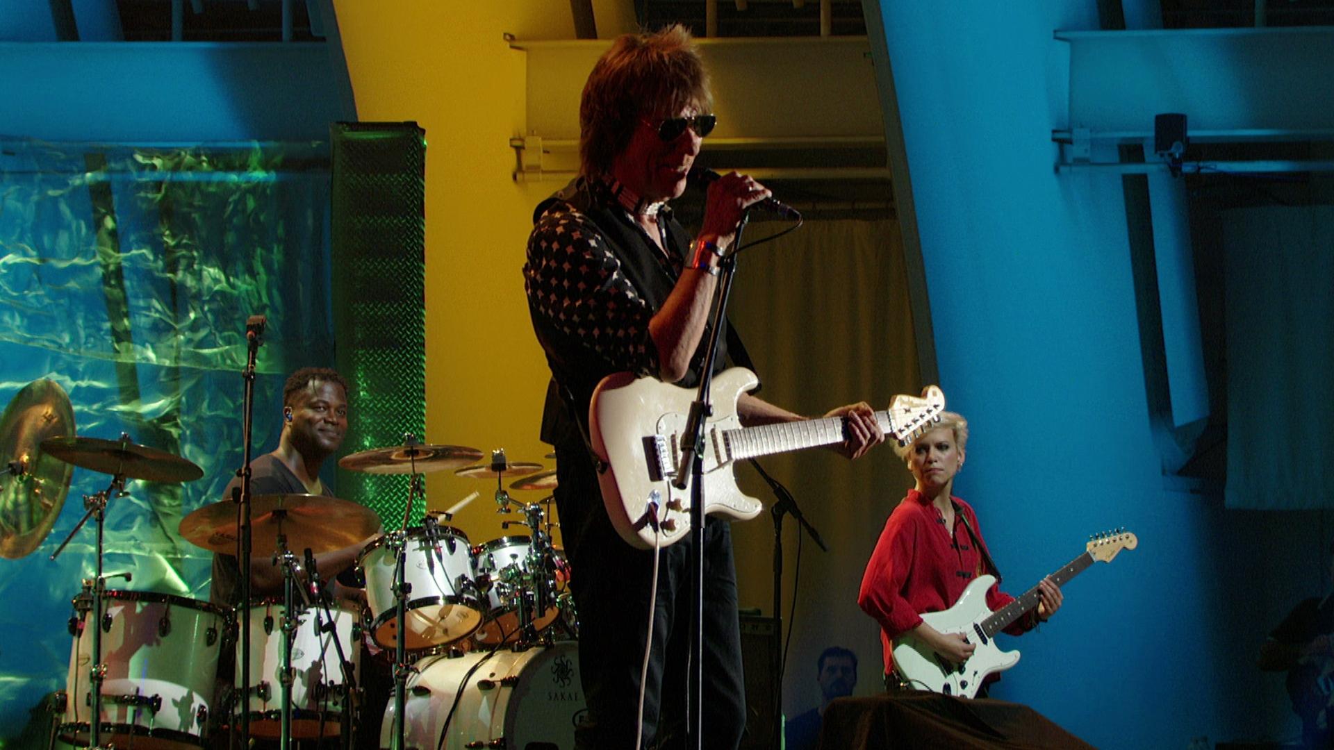 Jeff Beck performs onstage with a guitarist and lead singer in the center, a drummer to the left, and a bassist on the right.