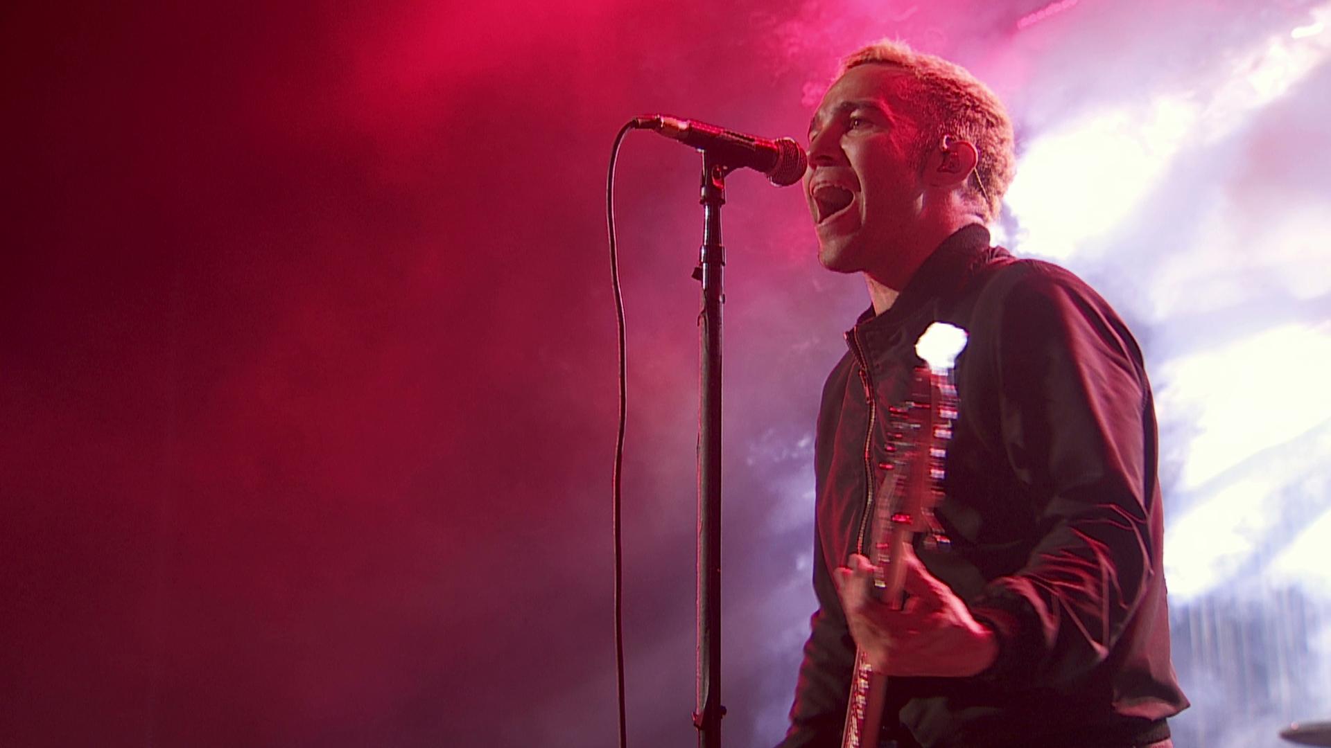 Fall Out Boy bassist singing into a microphone on stage with dim, red lighting and holding a guitar.