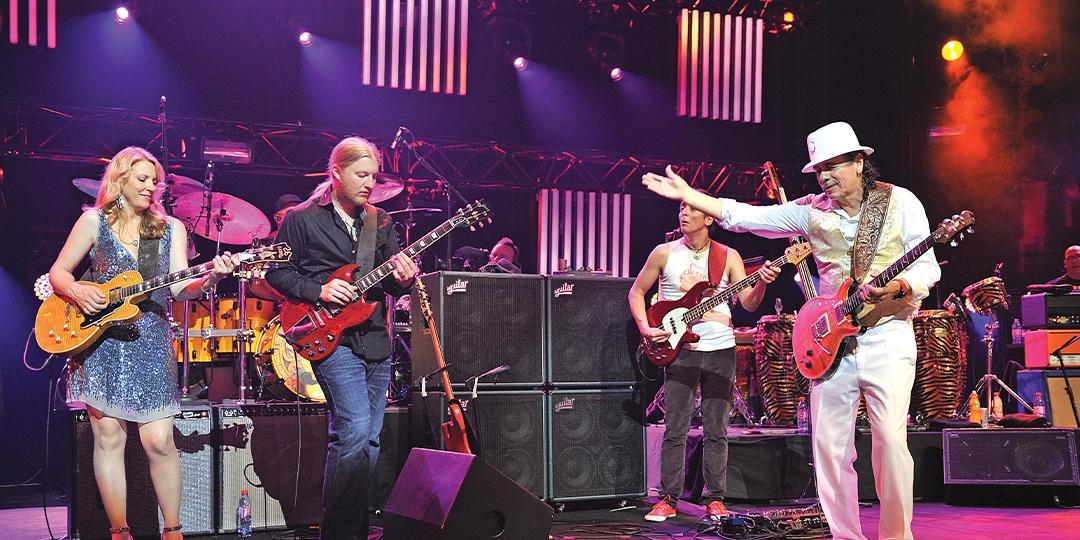 Santana perform on stage with guitars, bass, and drums. The scene is colorful with bright stage lights, and one performer in a white hat gestures with his arm extended.