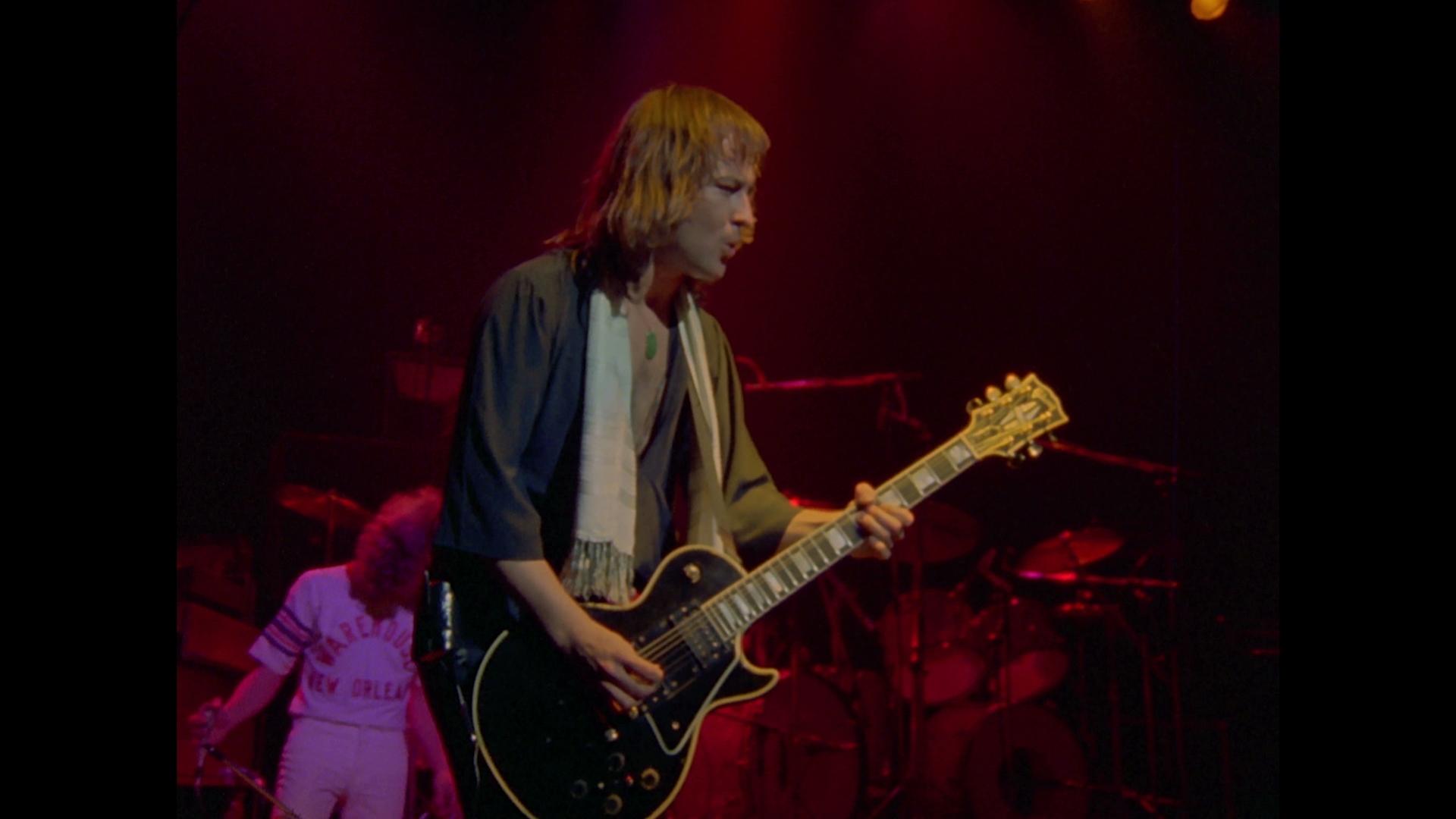 Foreigner guitarist, standing on stage under red lighting, plays an electric guitar while another band member in a white outfit stands in the background.