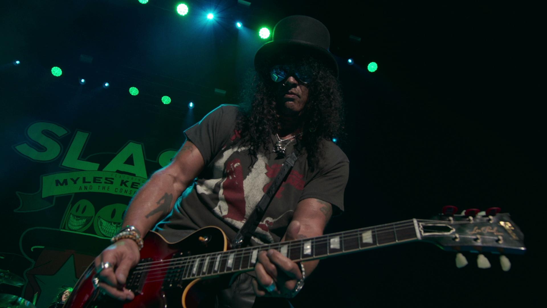 Slash wearing a top hat and sunglasses plays a red electric guitar on stage, with green spotlights in the background.