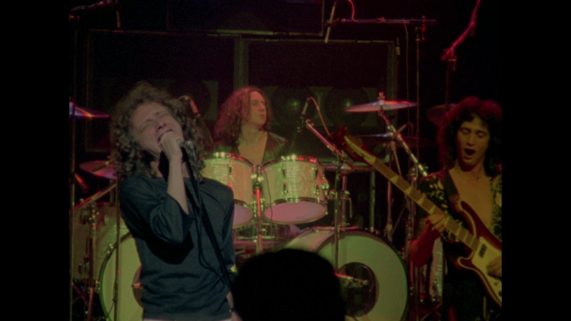 Foreigner performs on stage; the singer holds a microphone and sings passionately, with the drummer and guitarist playing in the background.