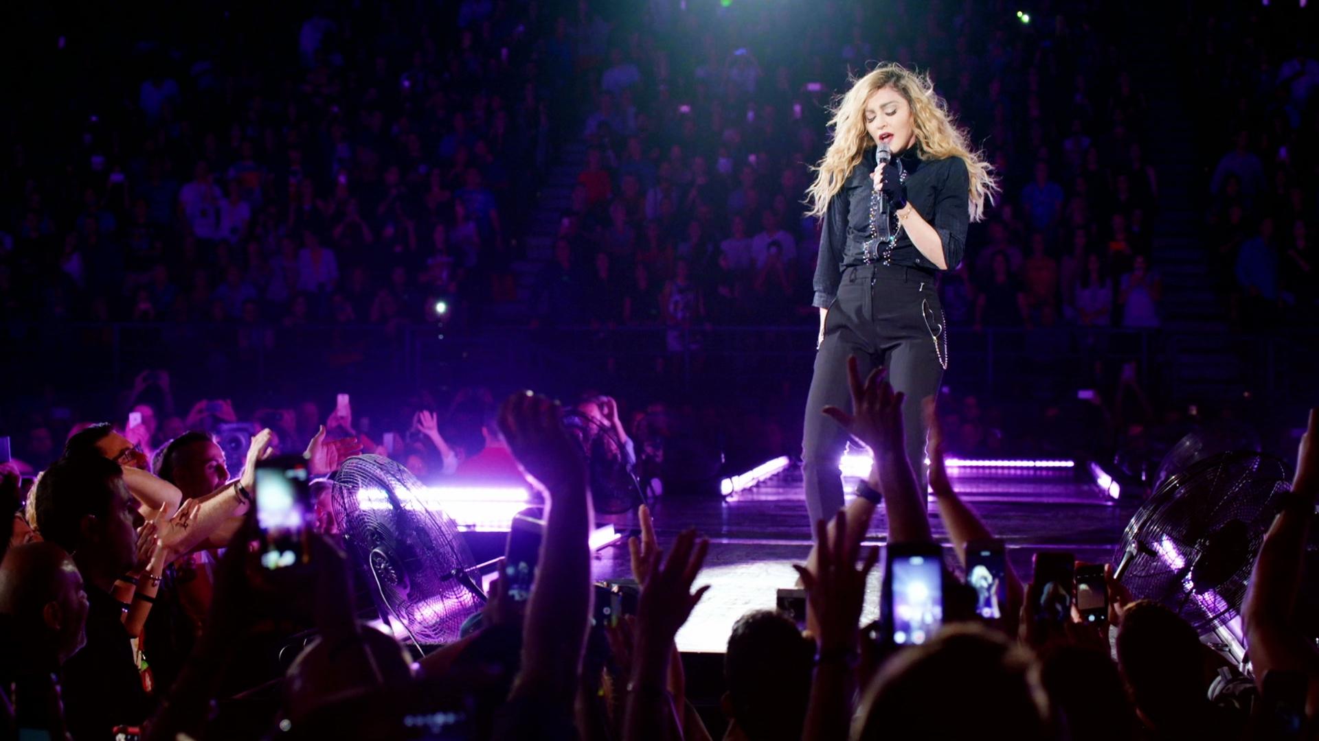 Madonna performs on stage at concert with crowd and bright lights surrounding her.