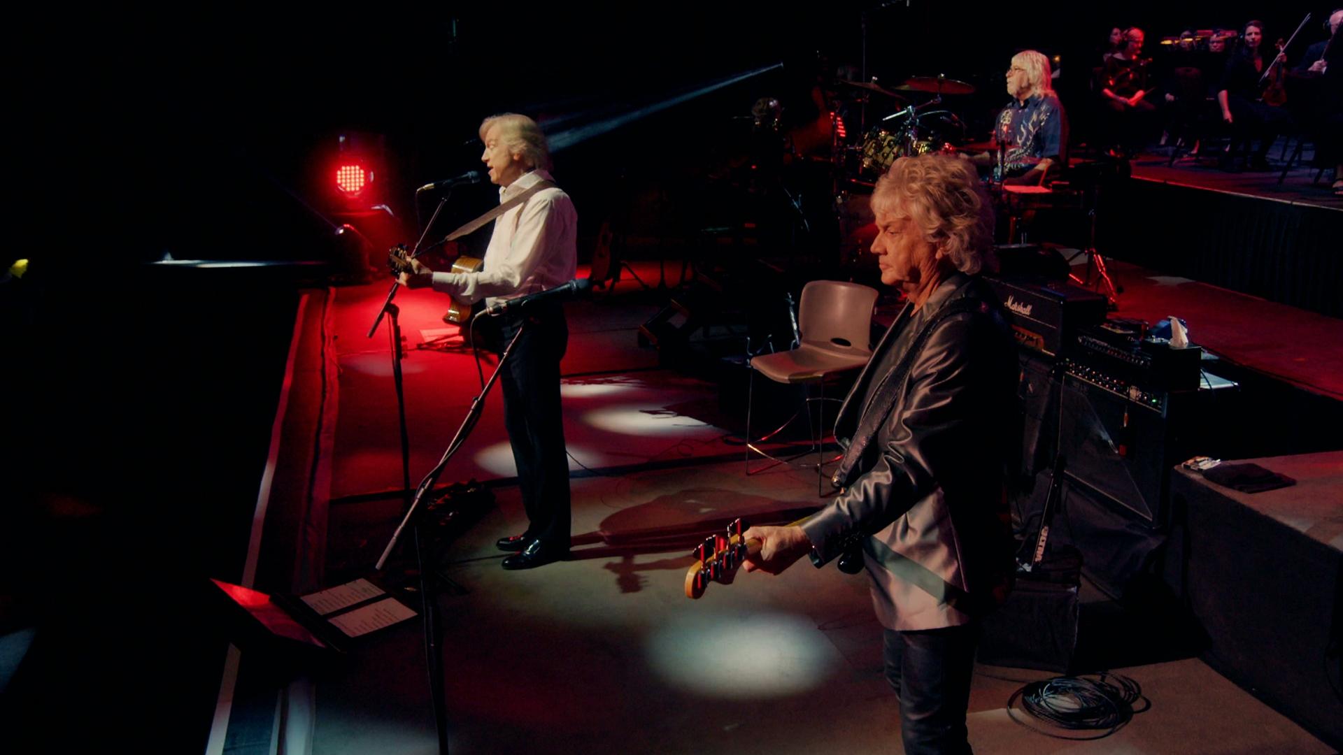 The Moody Blues perform on stage with guitars and drums, illuminated by red and spotlight lighting.