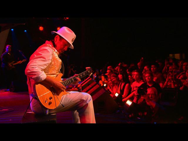 Santana in a white outfit and hat plays an electric guitar on stage with a crowd of people watching closely under red lighting.