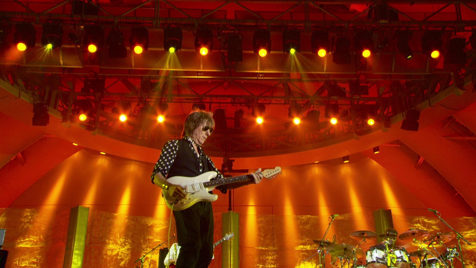 Jeff Beck stands on stage playing an electric guitar under bright orange and yellow lights. The stage is filled with lighting rigs and equipment.