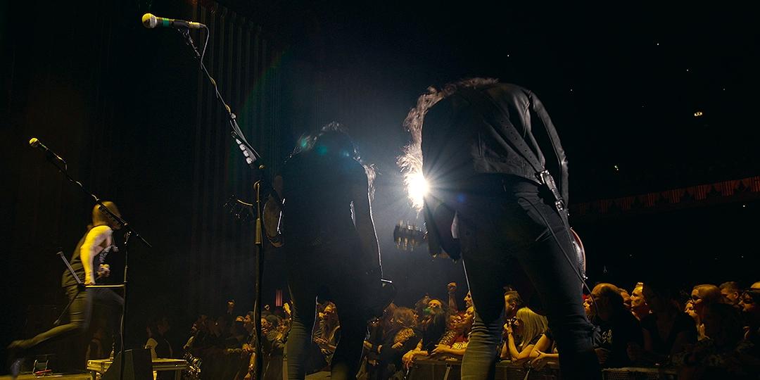 A band performs on stage with microphones and guitars under bright spotlights, facing a cheering audience.