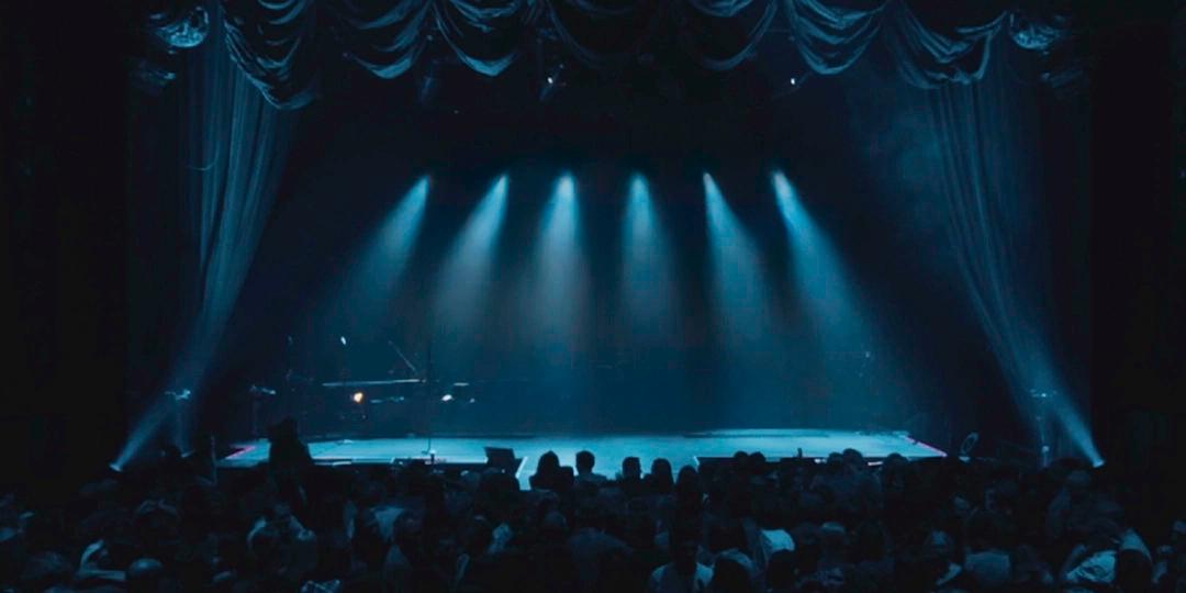 A dark stage is lit by six overhead spotlights pointing downwards. A piano is on the left side of the stage. An audience is seated in the foreground, facing the stage.