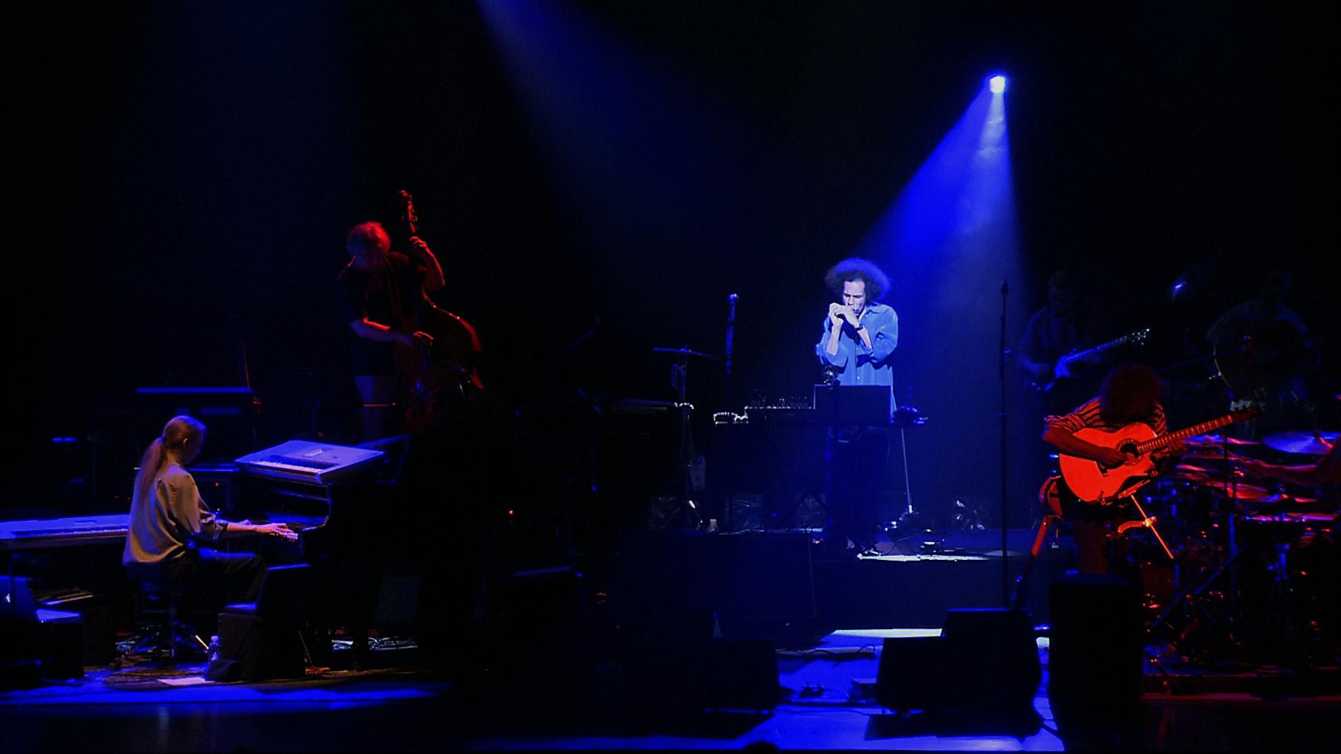 Pat Metheny performs on a dimly lit stage with blue and red lighting. A pianist, guitarist, singer, and other musicians are playing instruments.