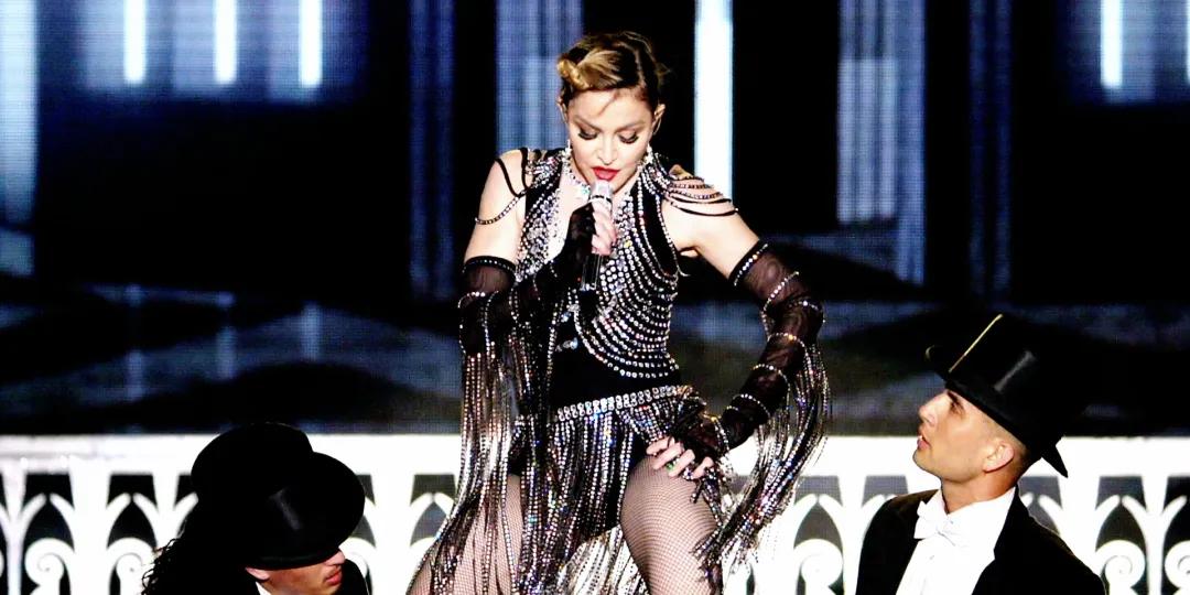 Madonna in a beaded outfit sings into a microphone on concert stage, flanked by two individuals in top hats.