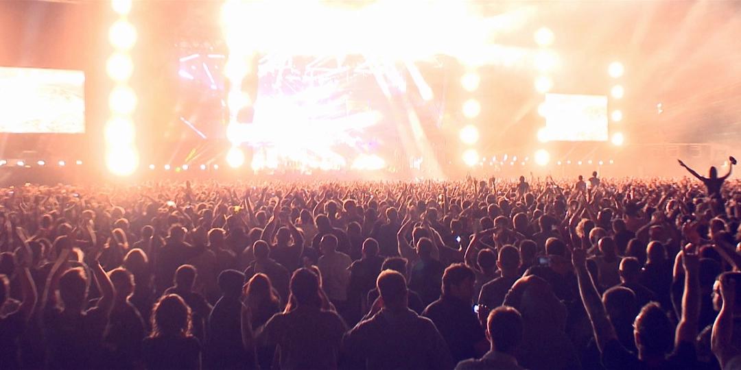 A large crowd at a Produgy concert with bright stage lights illuminating the scene. People are cheering and some are holding up their phones to capture the moment.