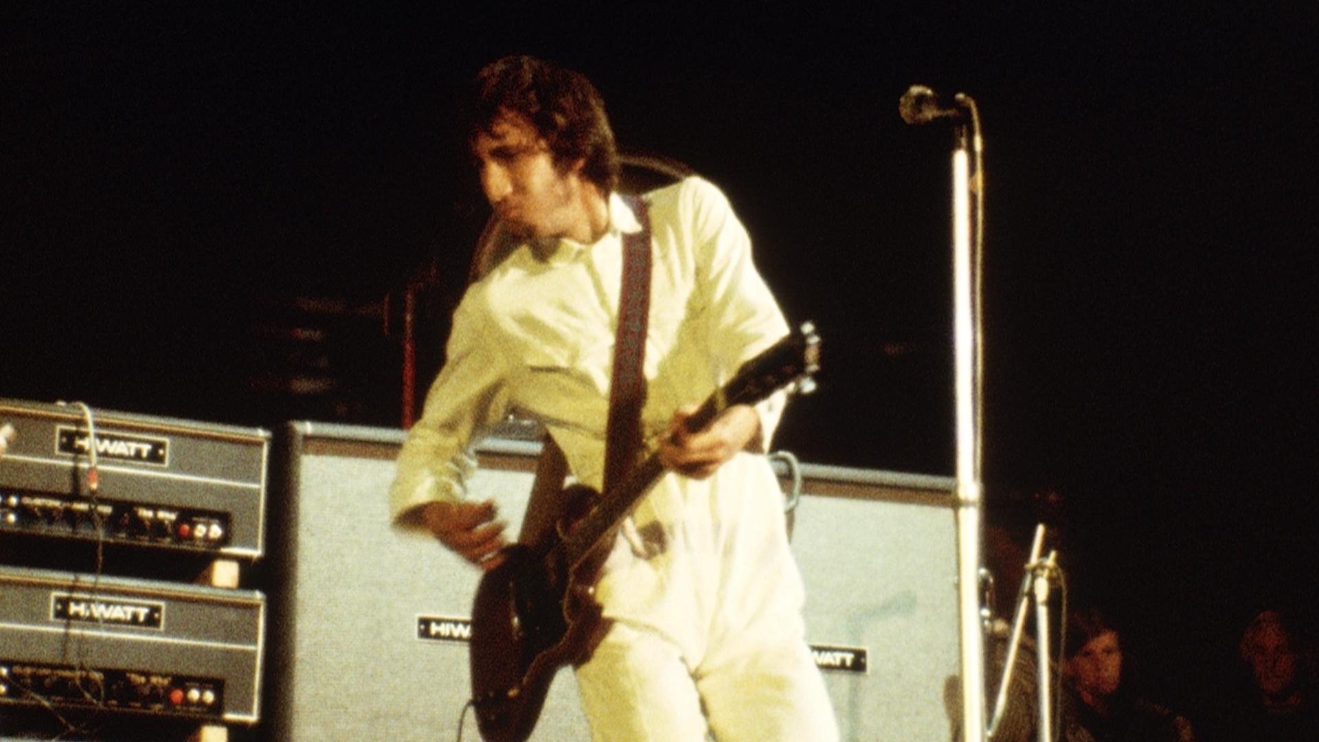 The Who performs on stage with a guitarist in a skeleton outfit, a vocalist with curly hair in the center, a drummer in the background, and a bassist on the right.