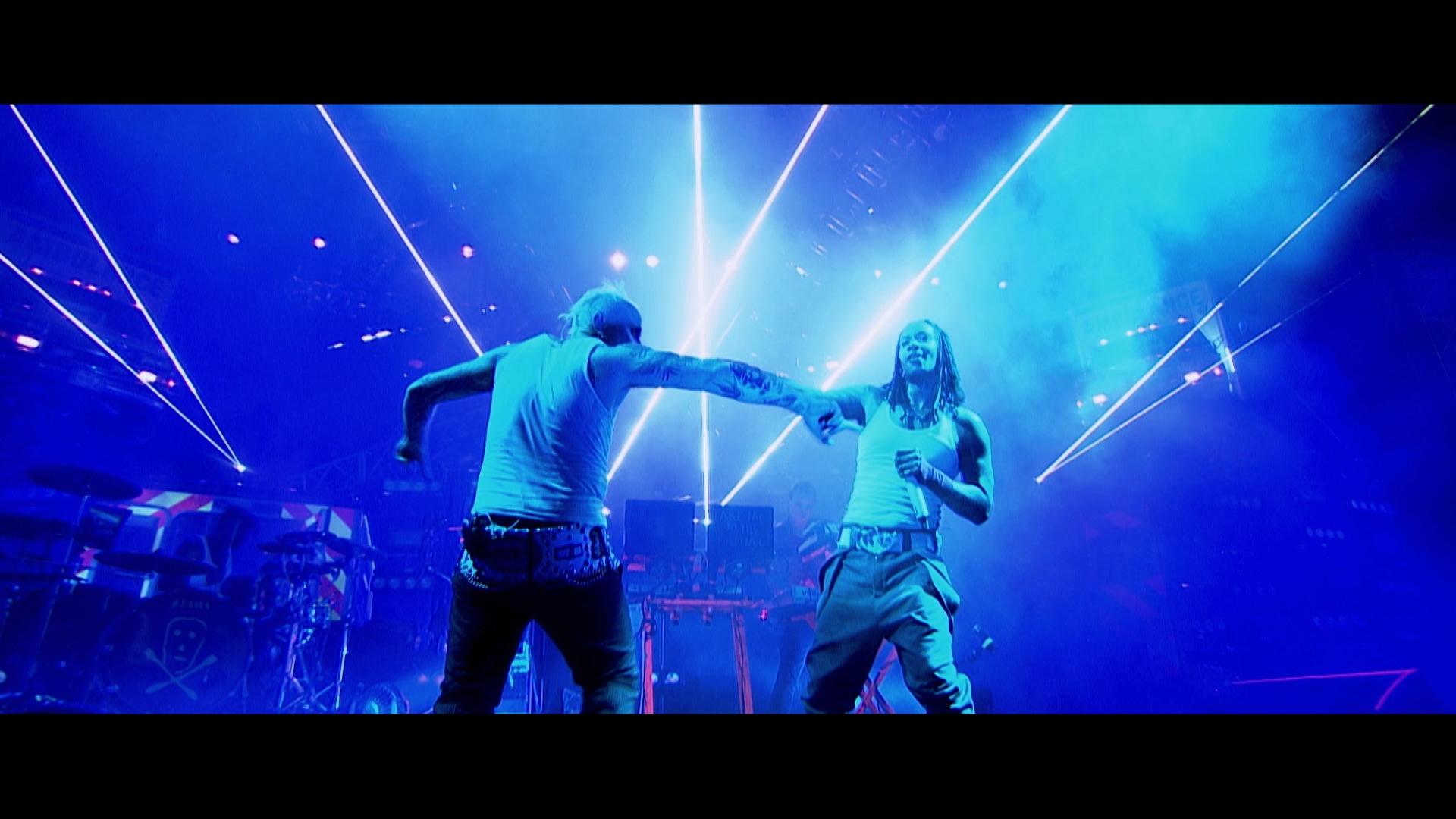 The Prodigy on stage, dressed in casual clothing, engage in a dynamic action with vibrant blue and purple stage lights and lasers illuminating the background.