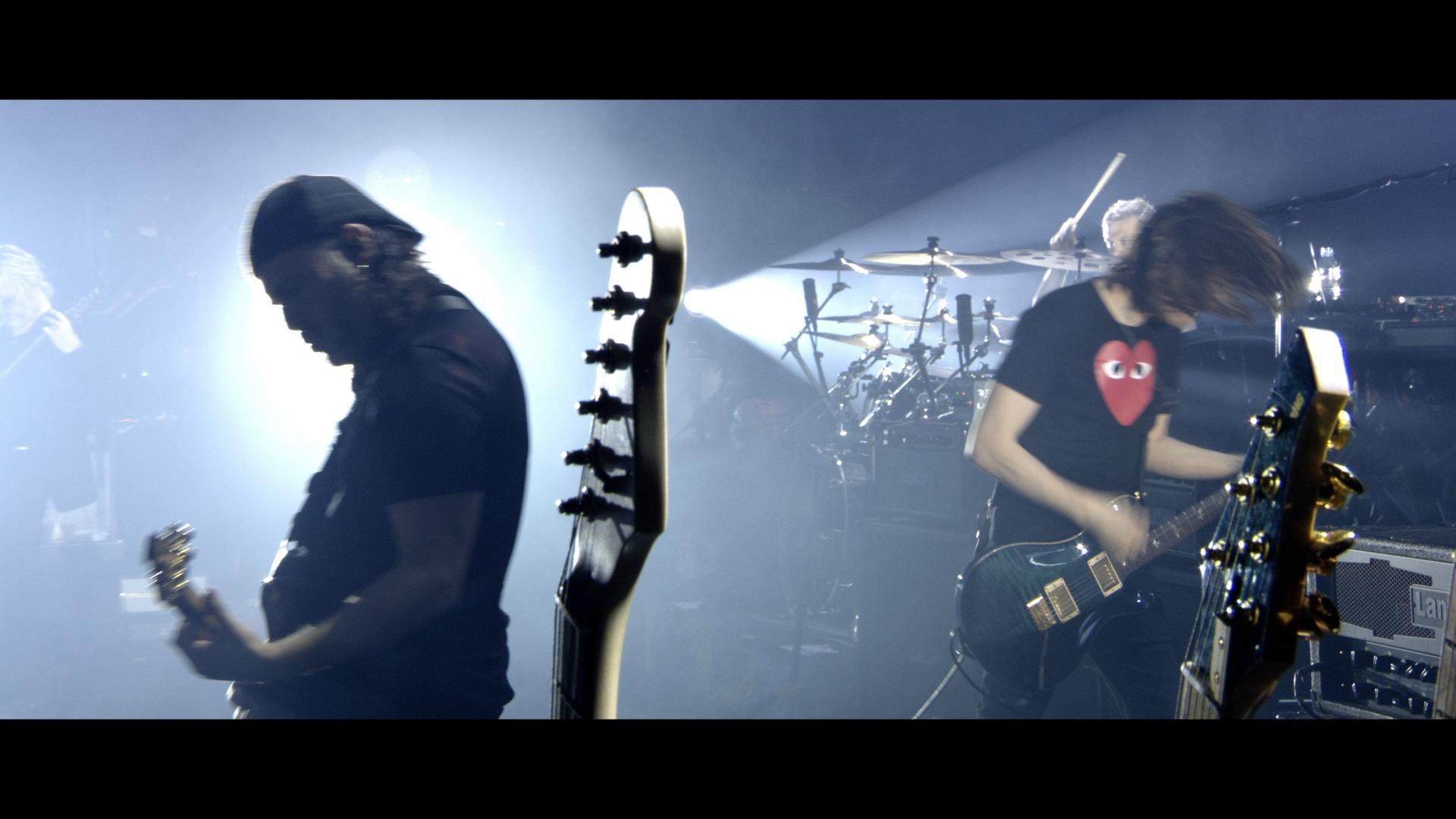 Steven Wilson performing on stage with intense lighting; one is in shadow wearing a cap and the other, with long hair, is illuminated, wearing a T-shirt with a red heart design. A drummer is visible in the background.