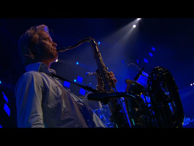 Simply Red band member on stage playing a saxophone under blue stage lights.