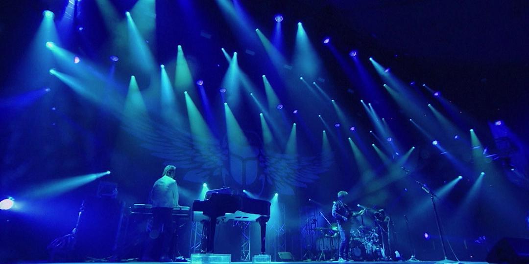 Journey performs on a stage with blue lighting and spotlights. The set includes a keyboard, a drum kit, and several guitars. A winged emblem is visible on the backdrop.