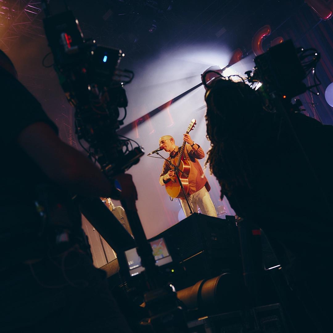 Bombay Bicycle Club frontman Jack playing an acoustic guitar on stage is illuminated by spotlight, with two camera operators filming the performance.