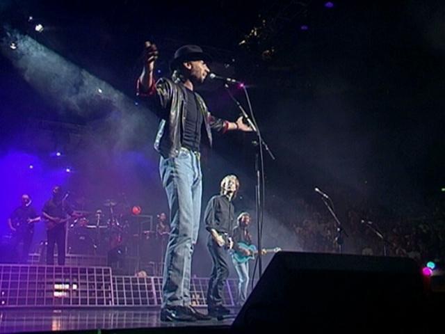 A Bee Gee in a black hat and leather jacket sings on stage with a microphone, while band members play instruments in the background under colorful stage lighting.