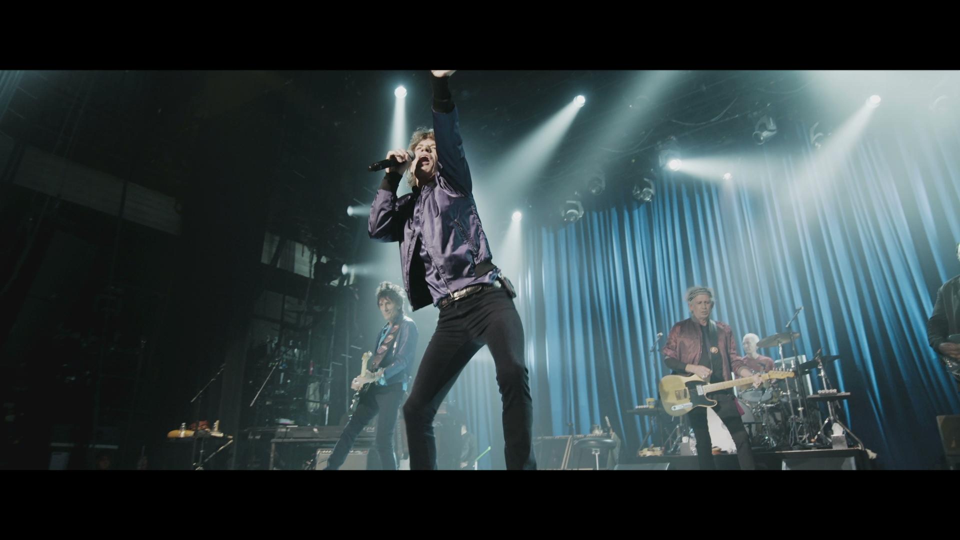 The Rolling Stones performing on stage under bright lights; the lead singer is in the forefront, reaching out with one arm, while the guitarist, bassist, and drummer play in the background.