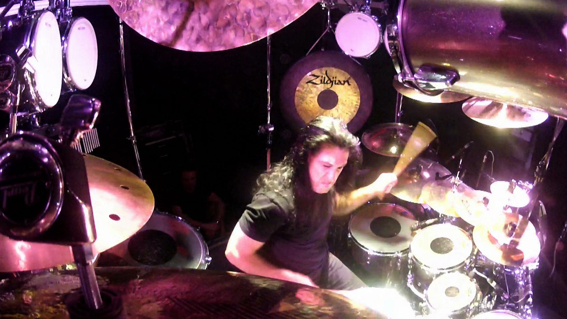 dream theater drummer with long dark hair is playing a drum set, surrounded by cymbals and drums, with a focused expression. The scene is well-lit and captured from above.