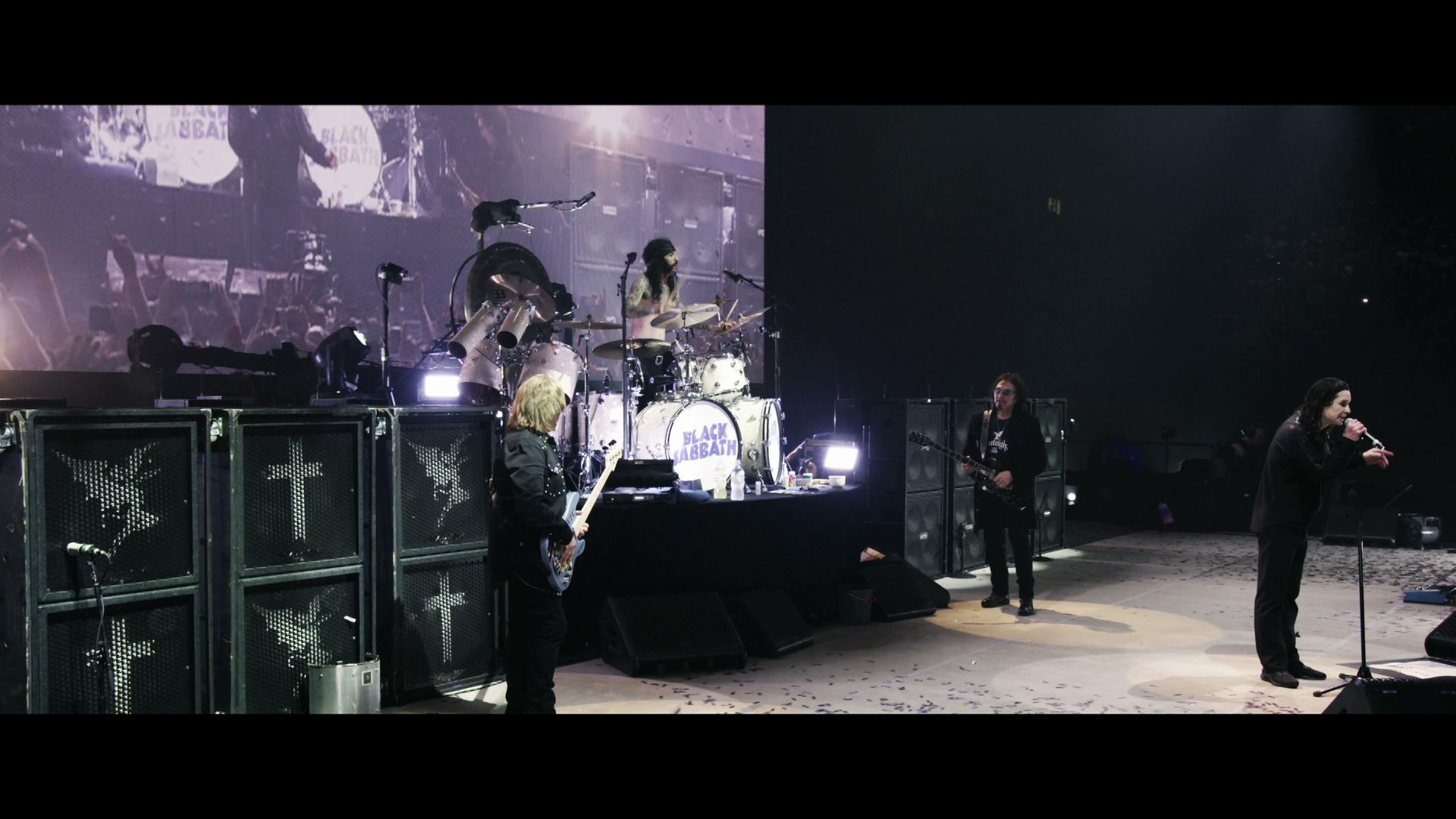 Black Sabbath performs on stage with one member singing, two playing guitars, and one on drums. A large screen in the background shows a live feed of the performance. Musical equipment and lighting are visible.