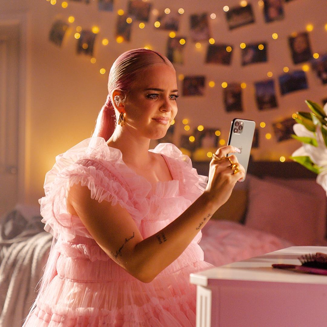 Anne-Marie with a slicked-back hairstyle in a pink, ruffled dress takes a selfie in a warmly lit room decorated with string lights and photos on the wall.