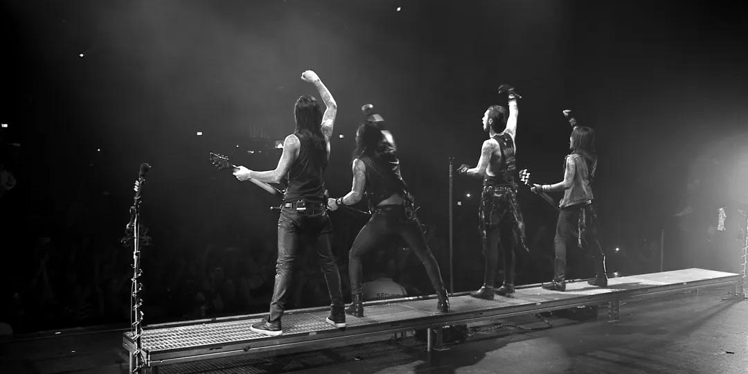 A black and white photo showing black veil brides performing on stage, facing away from the camera, with members raising their arms in front of a cheering crowd.