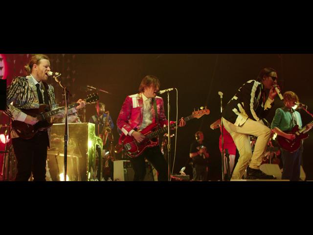 Arcade Fire performs energetically on stage with various musicians playing guitars and singing into microphones.