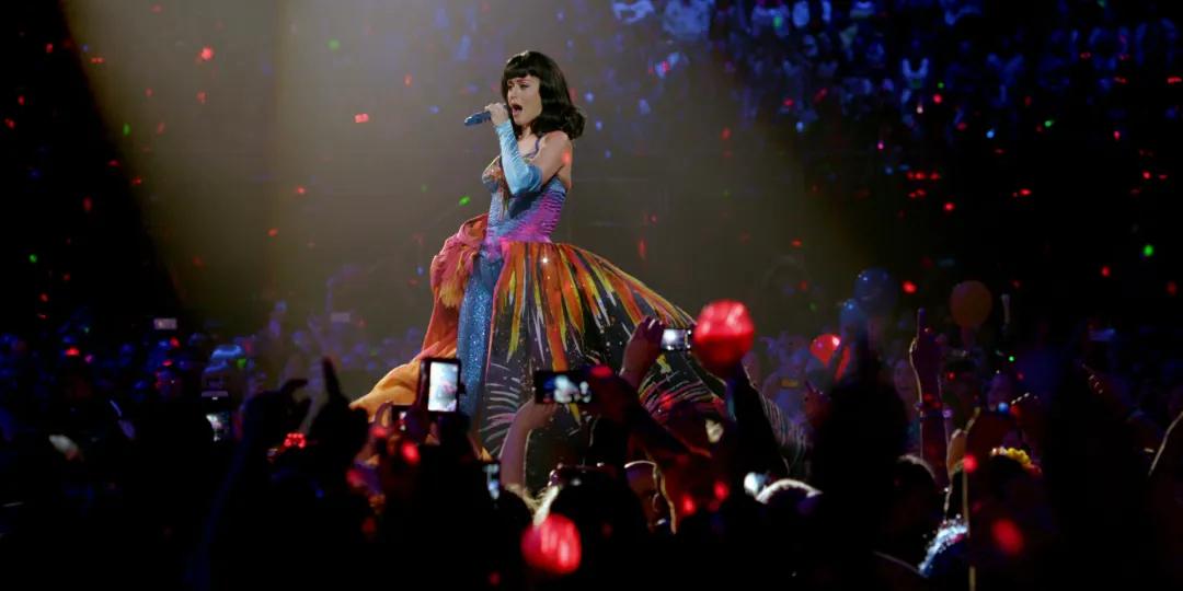 Katy Perry in a colorful dress sings into a microphone on a stage, surrounded by a crowd holding up lights and smartphones.