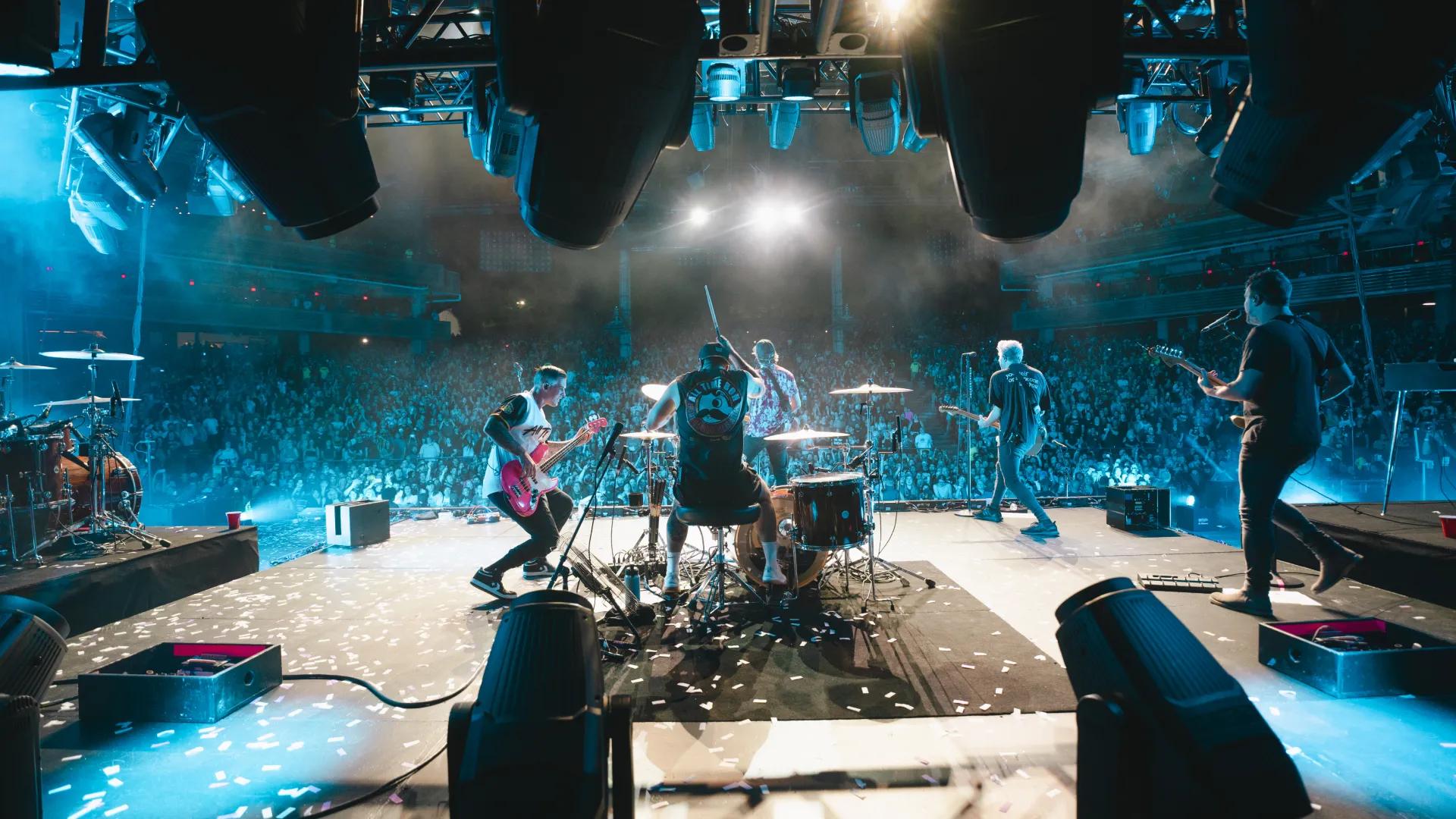 All Time Low performs on a stage in front of a large audience, with bright stage lights illuminating the scene. The drummer and guitarists are visible from the back, and the crowd is seen cheering.