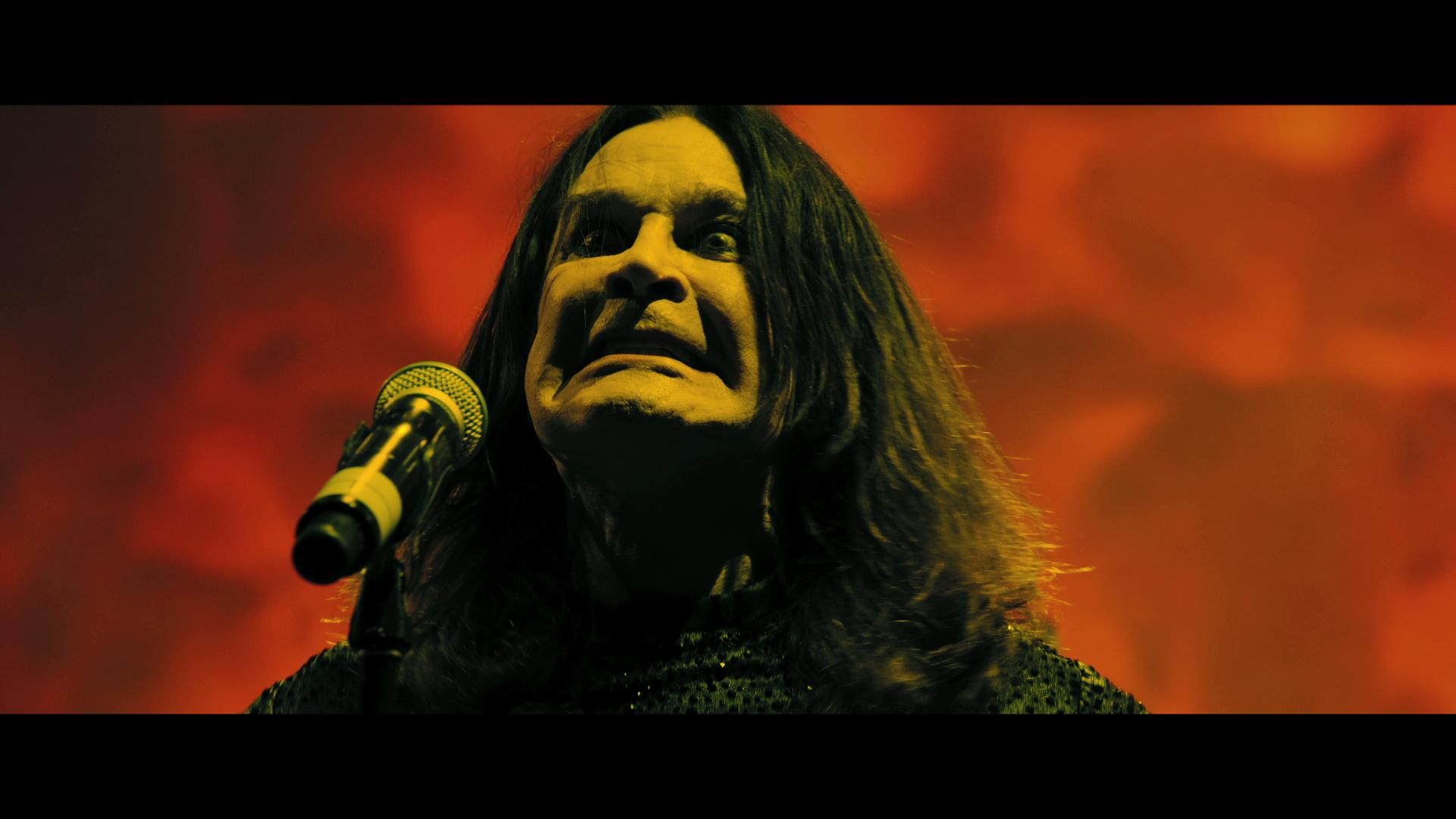 Ozzy Osborne with long hair, standing in front of a microphone, making a dramatic facial expression against a red and yellow background.