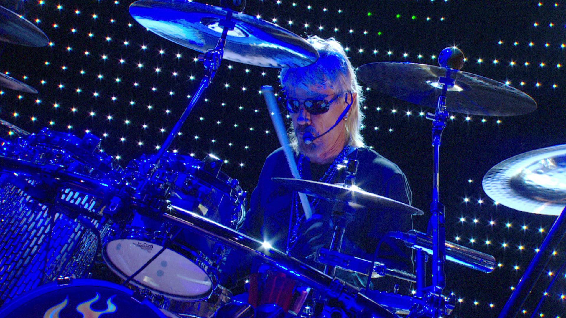 ZZ Top drummer wearing sunglasses plays a drum set on stage with bright lights in the background.