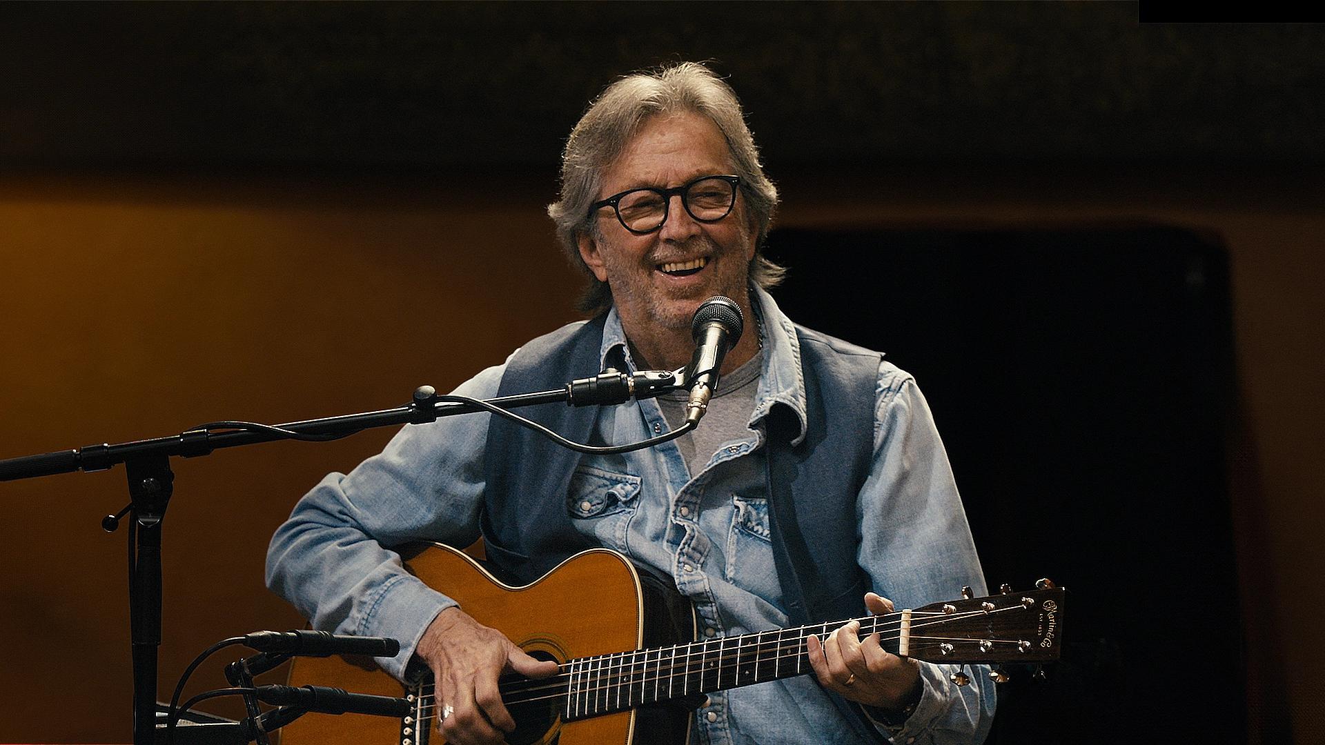 A man with gray hair and glasses, wearing a denim jacket, sits and plays an acoustic guitar while singing into a microphone.