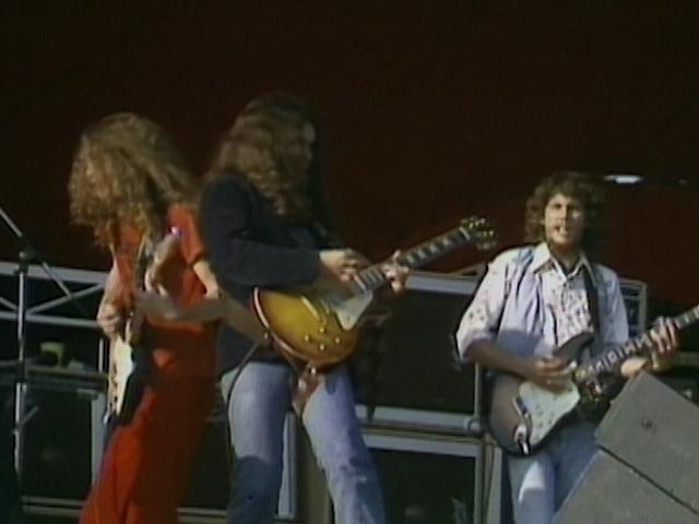 Lynyrd Skynyrd play electric guitars on stage, with amplifiers in the background. They have long hair and casually dressed in jeans and shirts.