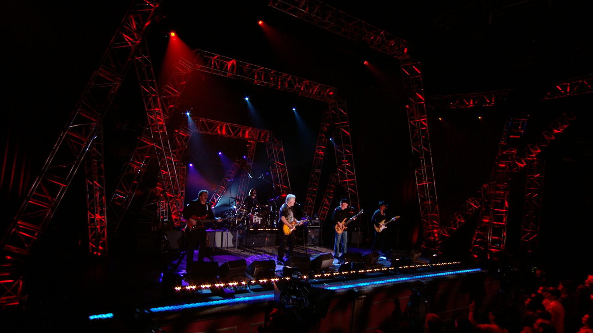 Bachman & Turner performs on a stage with red and blue lights, surrounded by metal scaffolding structures. The lead singer is playing a guitar.