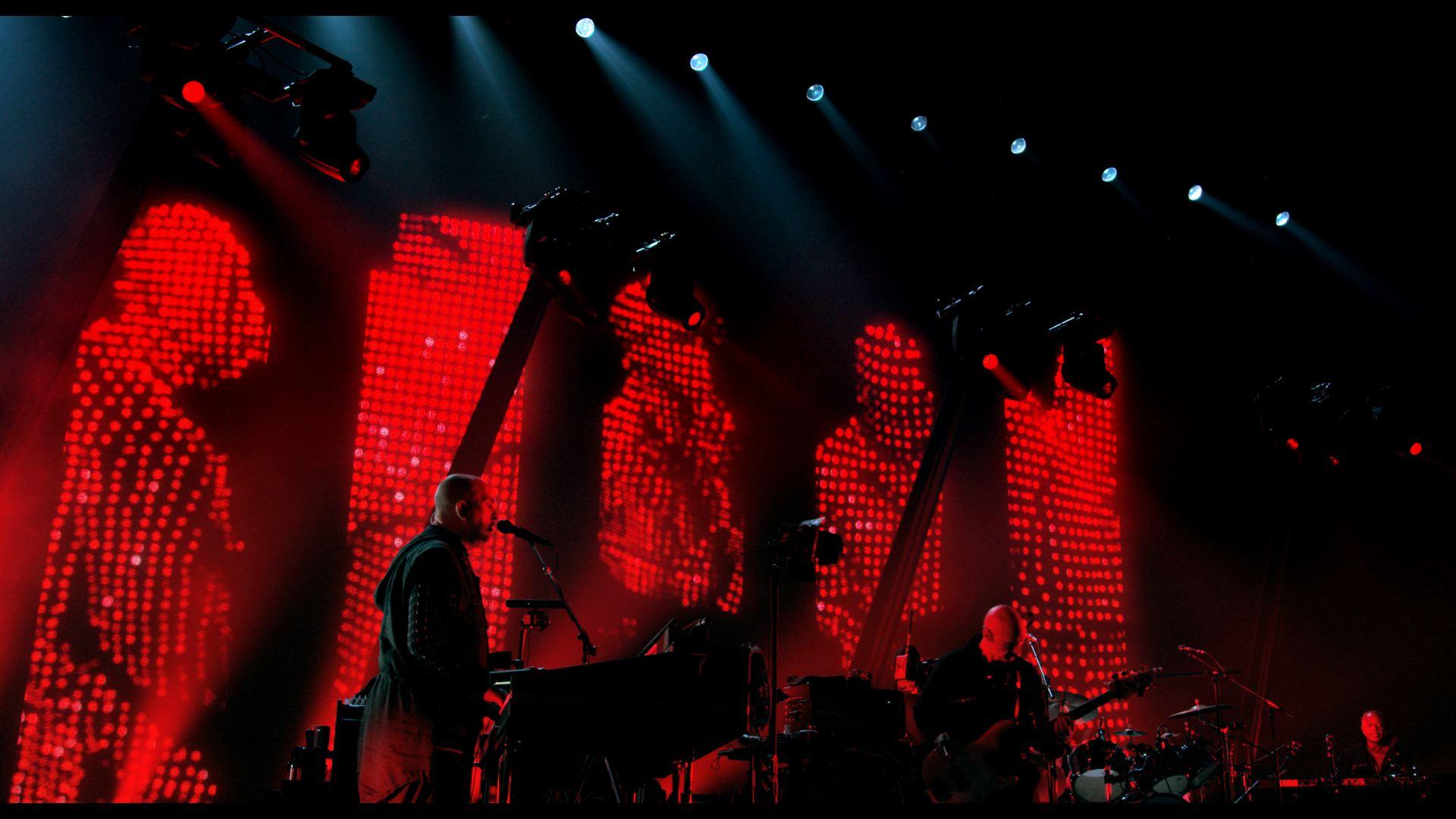 Peter Gabriel on stage under red lighting with digital screens displaying abstract figures in the background.