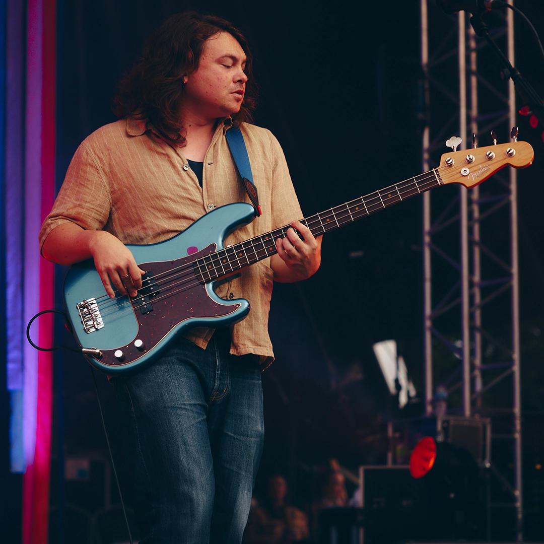 Bombay Bicycle Club bassist with long hair plays a blue electric bass guitar on stage, wearing a beige shirt and jeans. There is stage lighting with red and blue colors in the background.