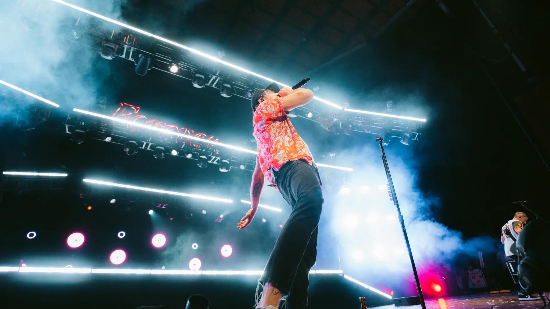 All Time Low frontman Alex on stage wearing an orange patterned shirt sings into a microphone, with bright stage lighting and smoke effects in the background.