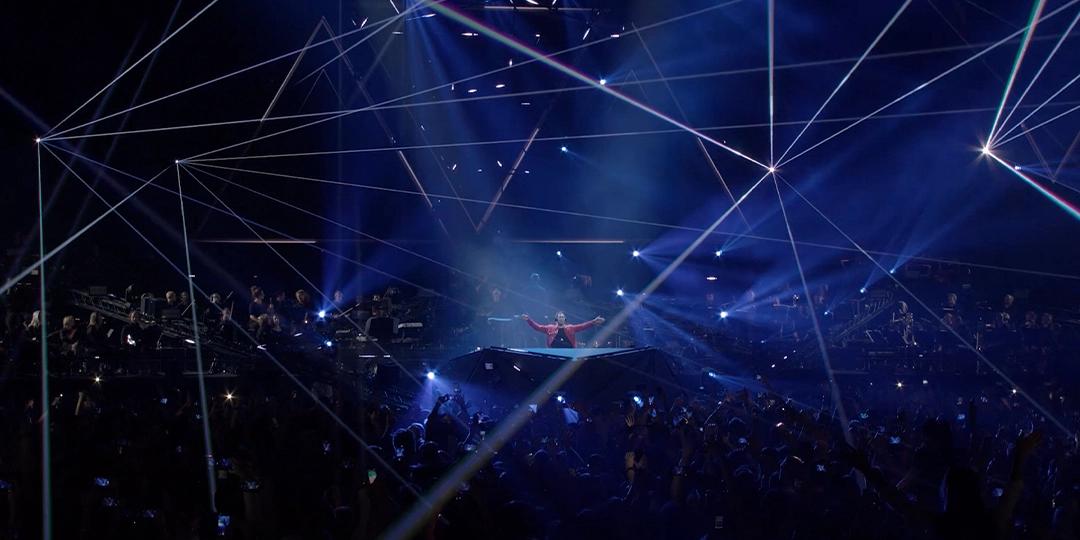 Hardwell performs on stage with a light show and an enthusiastic audience. Blue and white laser lights crisscross overhead.