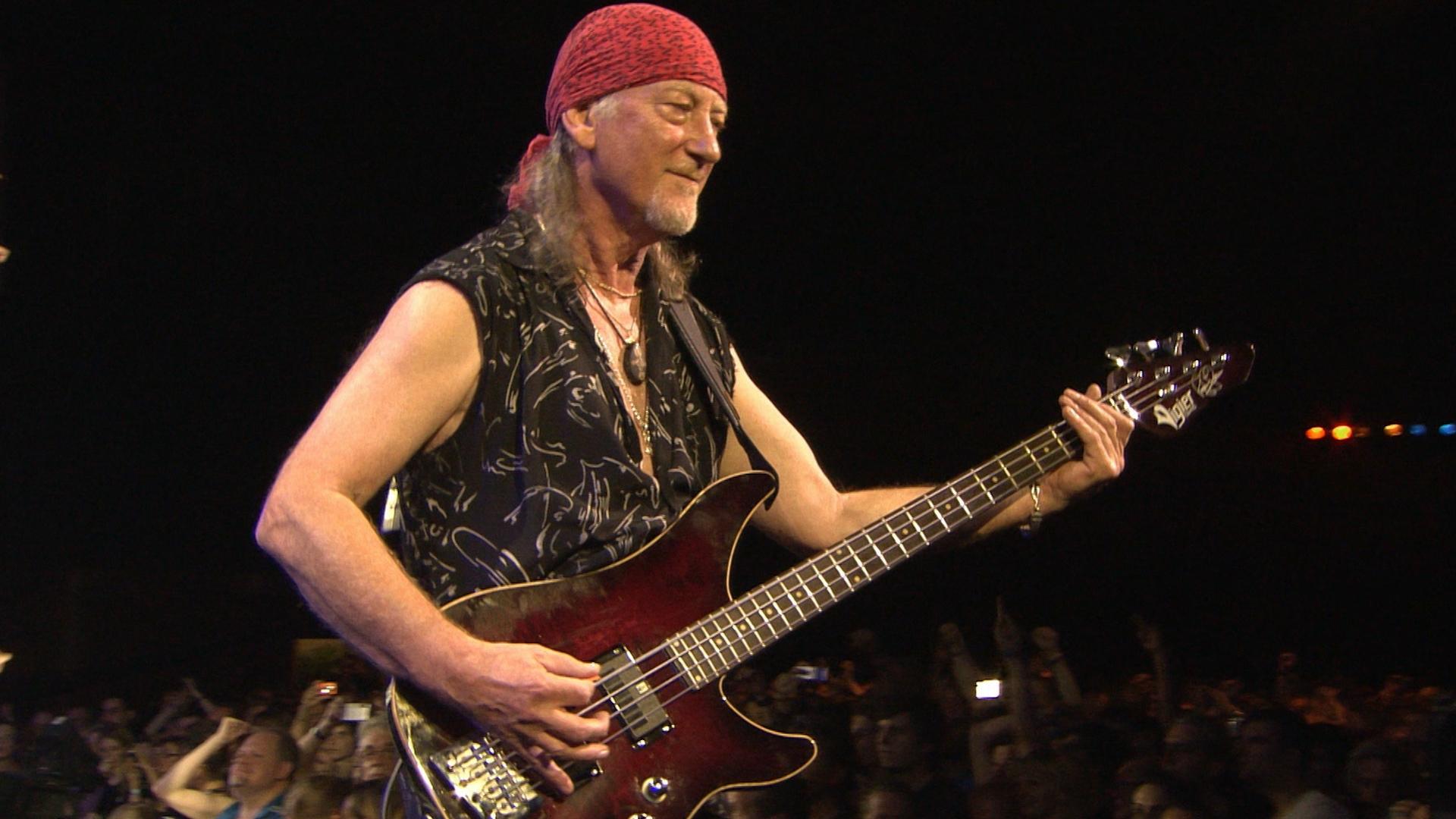 Deep Purple guitarist wearing a red bandana and sleeveless shirt plays an electric guitar on stage with an audience in the background.