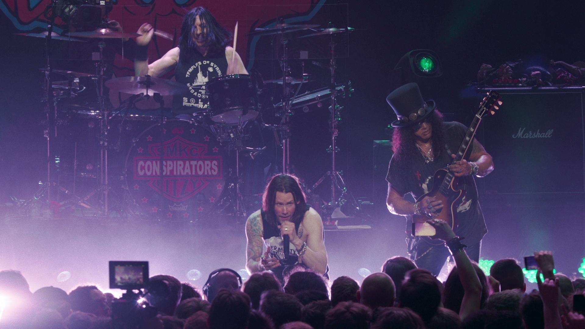 Slash featuring Myles Kennedy and the Conspirators perform on stage. The drummer is in the background, while the lead singer kneels at the front, and the guitarist with a top hat plays on the right. The audience is visible in the foreground.