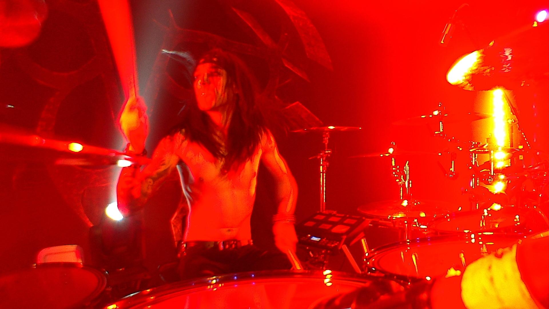 Black Veil Brides drummer with long hair plays energetically on a drum set, bathed in red stage lighting.