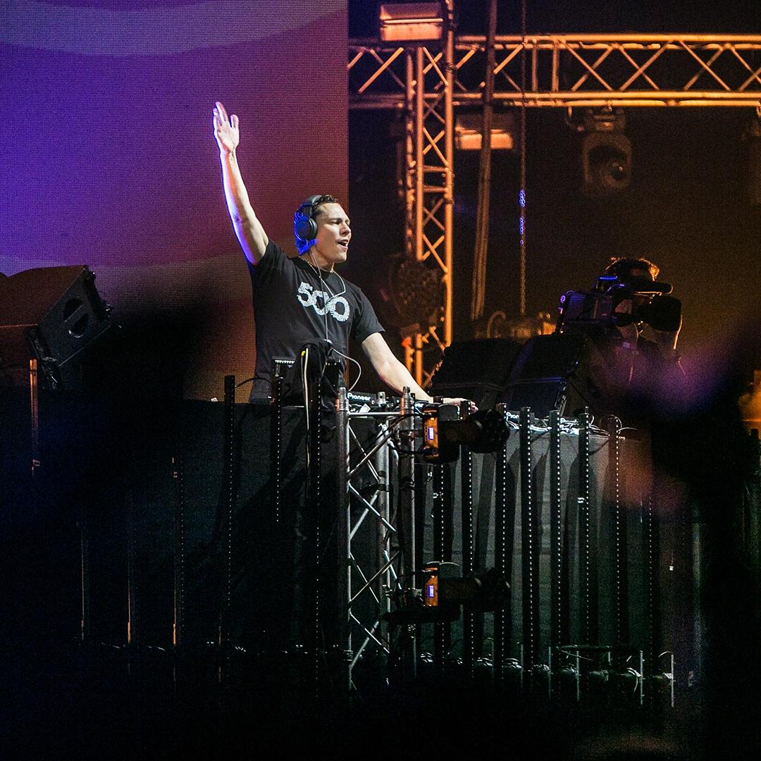 Tiësto wearing headphones is DJing on a stage, raising one hand while playing music, with lighting equipment visible in the background.