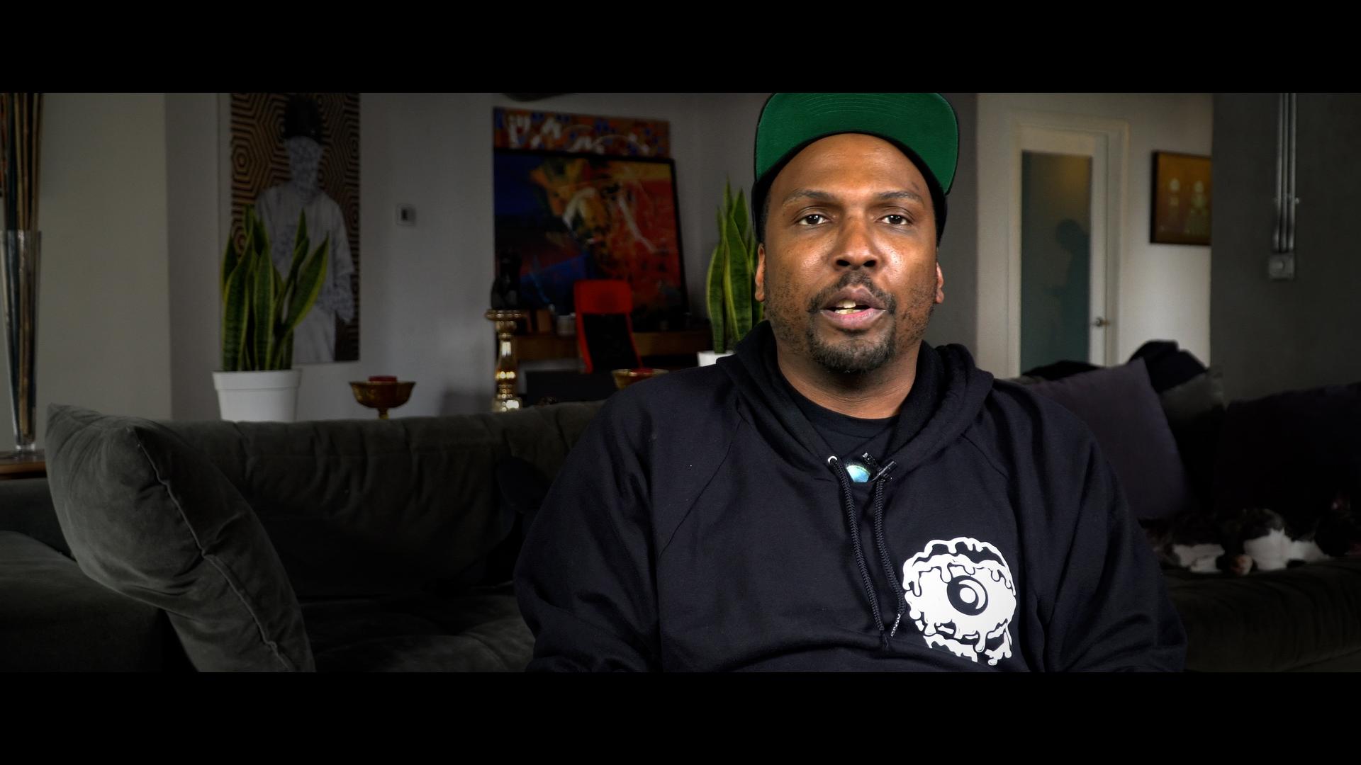 Bass Impact contributor wearing a black hoodie and green cap sits on a couch in a room with modern decor, including plants and artwork on the wall.