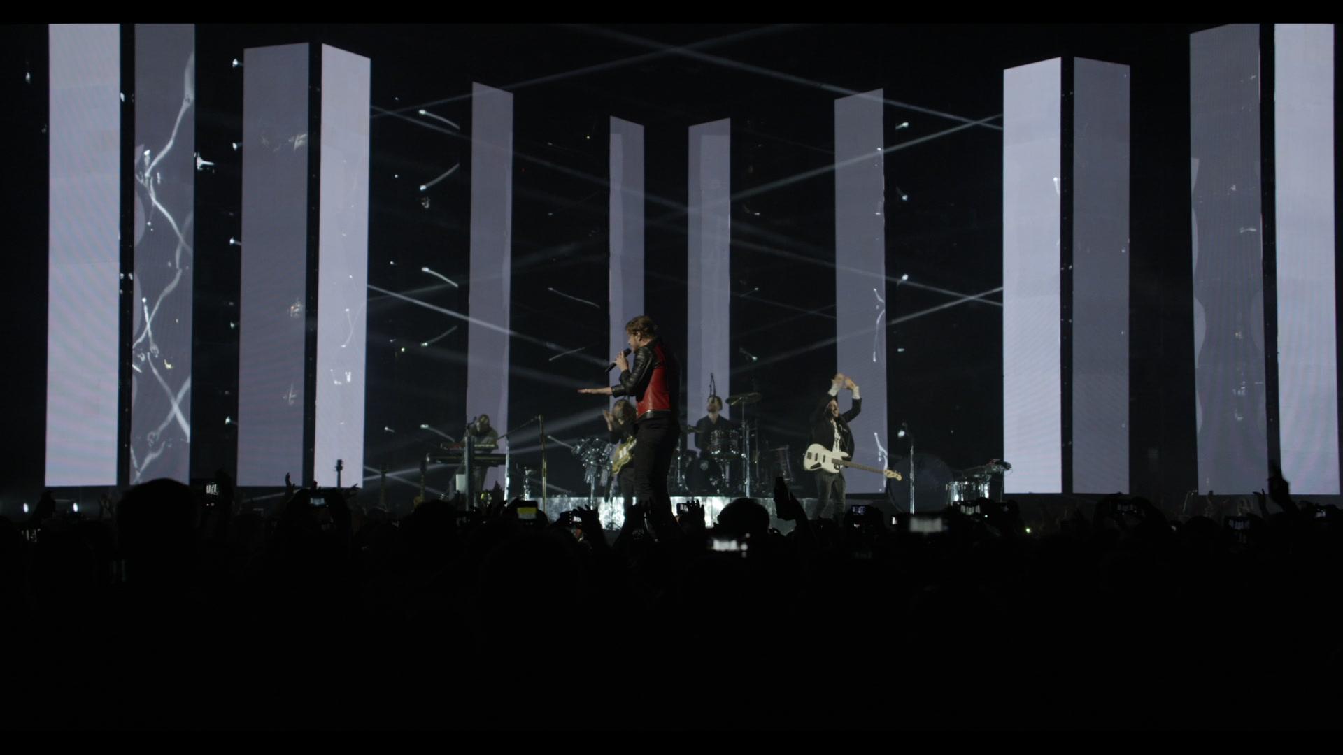 Imagine Dragons performs on stage with large vertical light panels and beams of light projected in various directions. The audience is visible in the foreground.