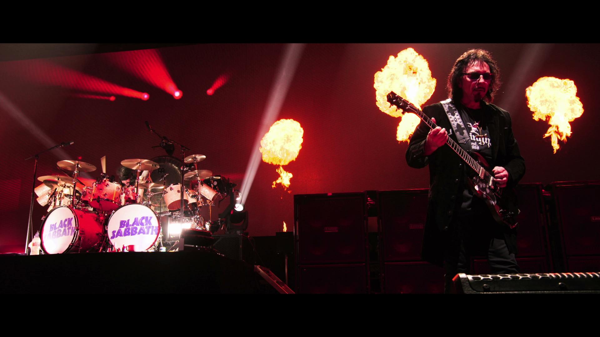 Black Sabbath perform on stage with flaming pyrotechnics behind them. The drum set has "Black Sabbath" written on it.