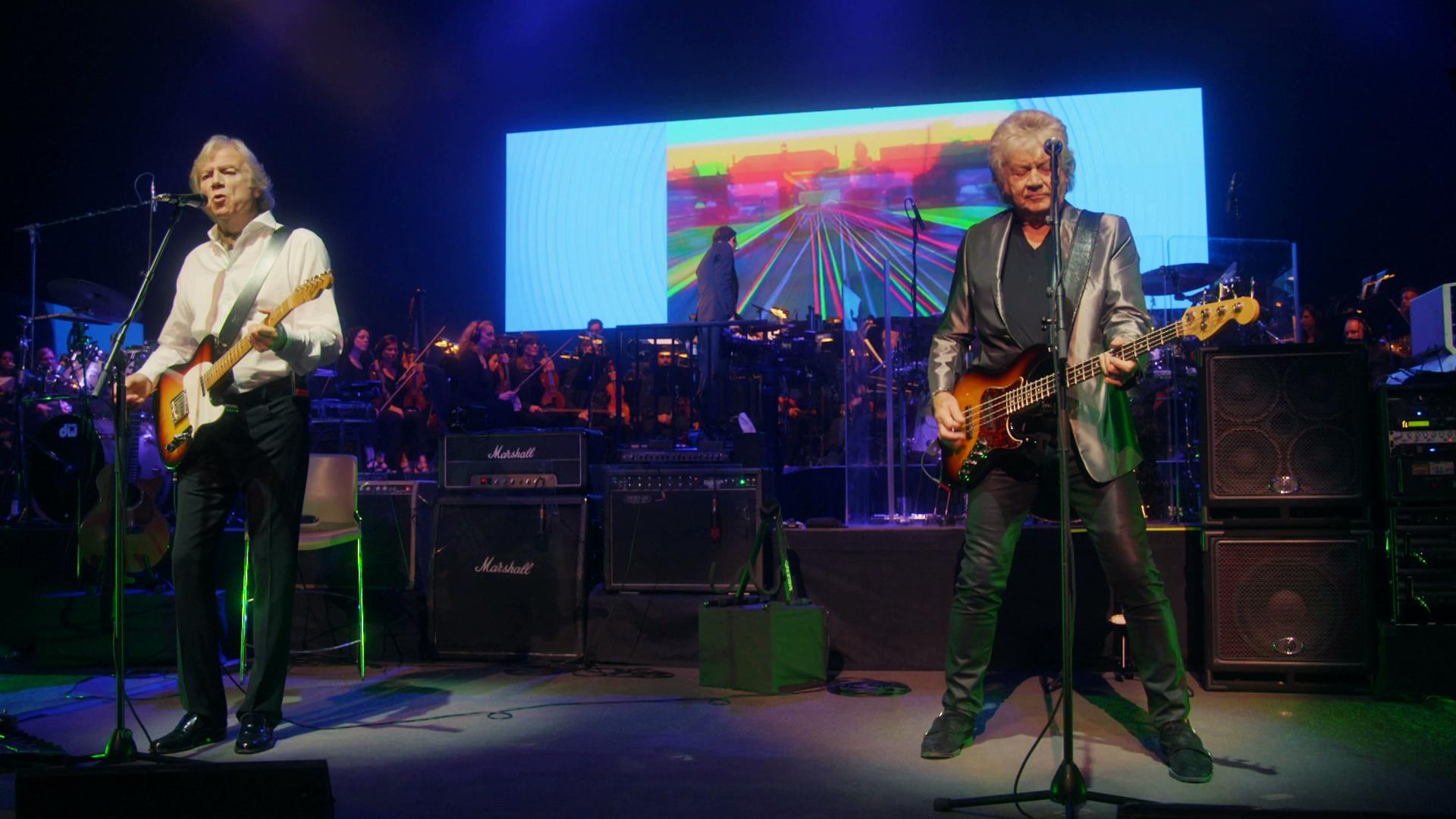 The Moody Blues perform on stage, one playing guitar and the other bass, with a colorful screen and orchestra in the background.